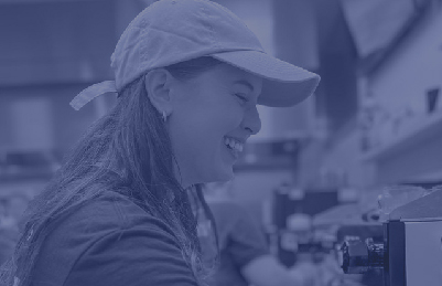 image of a cute young female barista with a smile on her face. shes wearing a hat and pouring a cup of coffee.